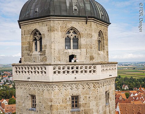 Steinerner Turm mit Rundbogenfenstern und Balustrade, Person mit Blasinstrument auf dem Balkon.