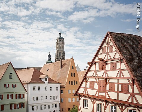 Fachwerkhäuser mit roten Dächern, im Hintergrund ein Kirchturm unter blauem Himmel.