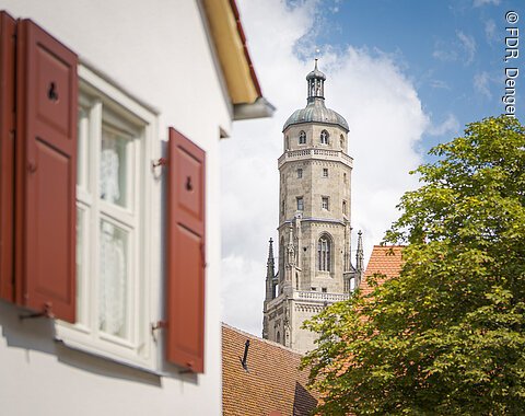 Ein Kirchturm ragt hinter einem Gebäude mit roten Fensterläden und einem Baum hervor.