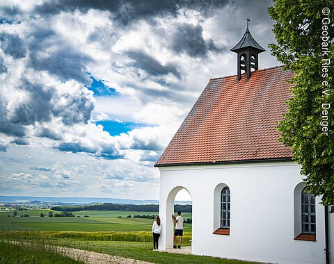 St. Antoniuskapelle bei Belzheim Kleine weiße Kapelle mit rotem Dach in ländlicher Umgebung, zwei Personen stehen davor.