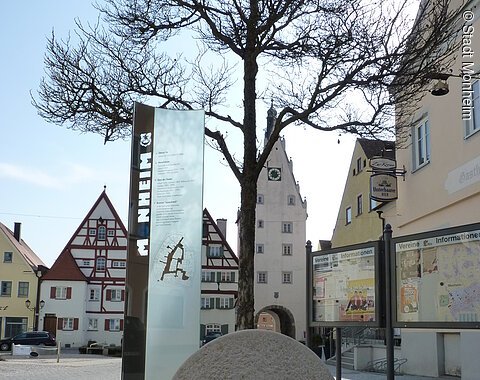 Buchstabenweg M-O-N-H-E-I-M Im Vordergrund ein großer grauer Buchstabe "O" aus Stein mit einer Infotafel daneben auf einem gepflasterten Platz. Dahinter, rechts im Bild zwei weitere Informationsvitrinen der Stadt Monheim, umgeben von historischen Gebäuden im Hintergrund. Der Buchstabe ist ein Teil des Monheimer Buchstabenweges.