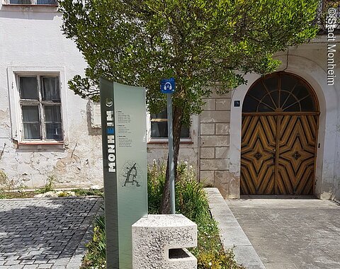 Buchstabenweg Monheim Steinskulptur des Buchstabens "E", dahinter ein Informationsschild und ein Baum mit einem dahinter liegendem Beet. Umgeben von einem gepflastertem Platz, mit einem historischen Gebäude im Hintergrund.