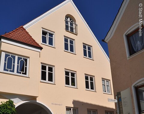 Buchstabenweg M-O-N-H-E-I-M Ein dreistöckiges Gebäude mit Giebel, einem Türbogen und mehreren Fenstern auf einem gepflastertem Platz. Vor dem Gebäude steht ein Informationsschild und ein großer grauer Buchstabe "M" aus Stein. Der Buchstabe ist ein Teil des Monheimer Buchstabenweges.