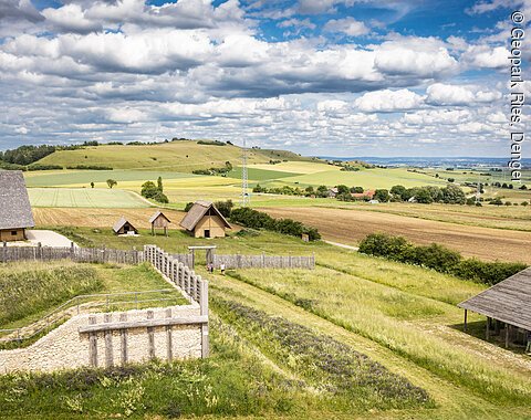 Landschaft mit Hügeln, Feldern und mehreren strohgedeckten Gebäuden, umgeben von einem Holzzaun.