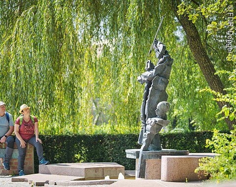 Zwei Personen sitzen auf einer Mauer neben einer abstrakten Skulptur im Freien, umgeben von Bäumen.