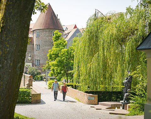 Zwei Personen spazieren auf einem gepflasterten Weg neben einer Backsteinmauer. Im Hintergrund ein Turm und Bäume.