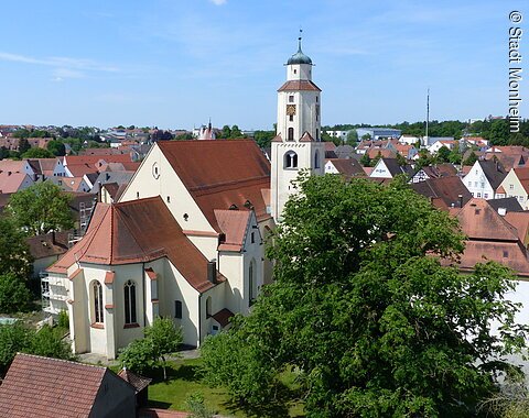 Luftbild der Stadtpfarrkirche mit Turm in einer Stadtlandschaft, umgeben von Häusern mit roten Dächern und Bäumen. Im Hintergrund blauer Himmel.
