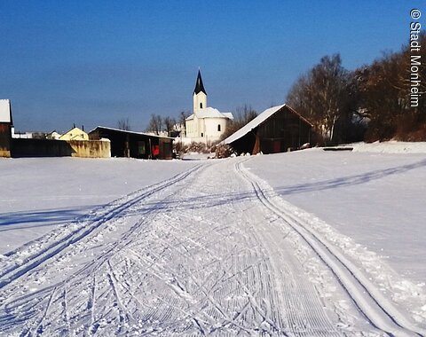 Verschneite Landschaft mit Skispuren, ein Ort zum Langlaufen in der Monheimer Alb. Im Hintergrund Gebäude, eine Kirche und blauer Himmel zu sehen.