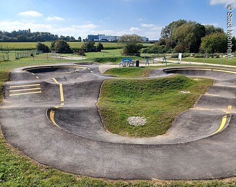 Pumptrack Monheim Asphaltierter Pumptrack mit gelben Markierungen in der Mitte, umgeben von grüner Landschaft und Bäumen im Hintergrund.