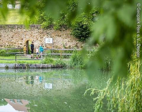 Klosterweiher, Kirchheim am Ries Eine Familie spaziert an einem Teich entlang, im Hintergrund eine Steinmauer und Bäume.