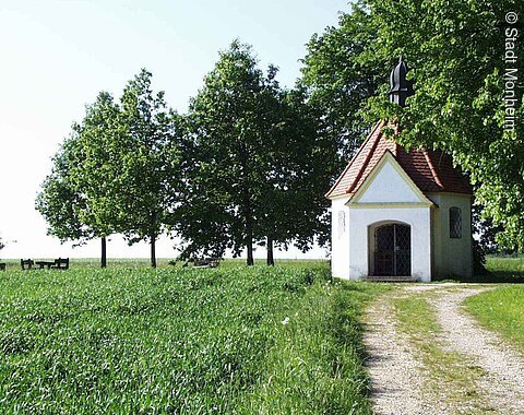 Kleine Kapelle mit rotem Dach auf einer Wiese mit einem Schotterweg. Umgeben von einem grünem Feld und Bäumen.