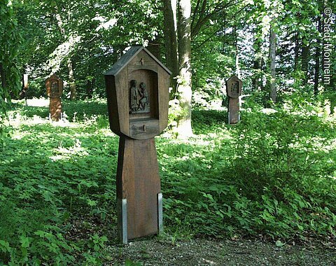 Holztafeln auf einem Kreuzweg mit religiösen Schnitzereien in einem bewaldeten Gebiet.