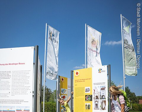 Ein Kind und eine Frau betrachten Informationstafeln im Freien, umgeben von Fahnen.