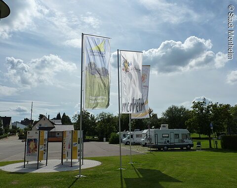 Drei Fahnen wehen im Wind, daneben stehen Informationstafeln auf einer Wiese. Im Hintergrund sind Wohnmobile und Bäume zu sehen.
