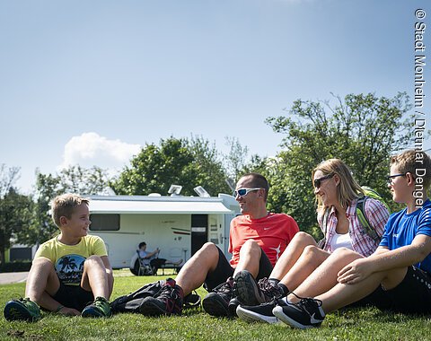 Eine Gruppe von vier Personen sitzt auf einer Wiese vor einem Wohnmobil und lacht.