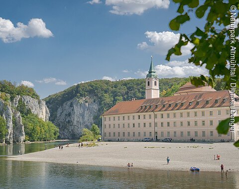 Flussufer mit Kiesstrand, altes Gebäude mit Turm und bewaldete Felsen unter blauem Himmel mit Wolken