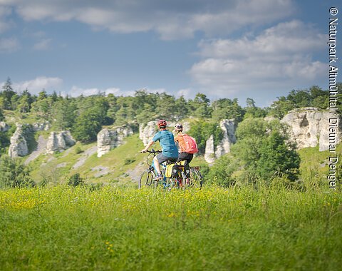 Zwei Personen fahren mit Fahrrädern auf einem grasbewachsenen Feld vor einer Felslandschaft.