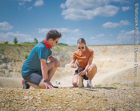 Zwei Personen knien auf einem steinigen Gelände und untersuchen den Boden.