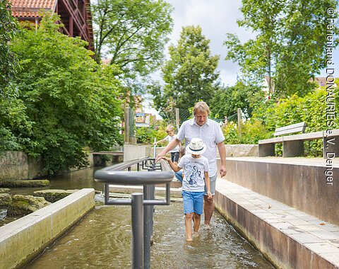 Ein Mann und ein Kind gehen barfuß durch ein Kneippbecken mit Wasser, umgeben von Bäumen und Bänken.