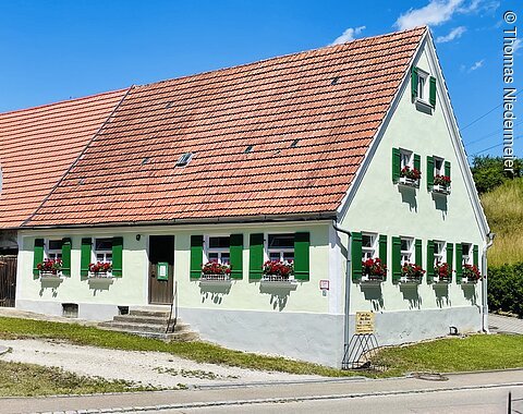 Weißes Haus mit grün lackierten Fensterläden und roten Blumen in Blumenkästen unter einem roten Ziegeldach bei blauem Himmel.