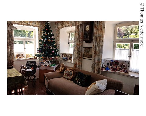 Wohnzimmer mit Weihnachtsbaum, Sofa, Puppen, Puppenwagen und Fenstern mit Gardinen und Blick ins Grüne