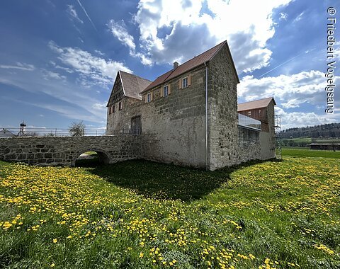 Altes Steingebäude mit rotem Dach auf einer Wiese mit gelben Blumen unter blauem Himmel mit Wolken.