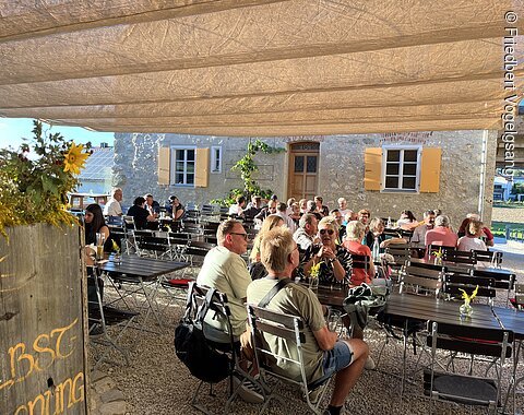 Sonnendurchflutete Terrasse mit Gästen an Tischen vor einem Haus mit gelben Fensterläden und Sonnensegeln.