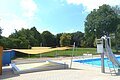 Freibad Monheim - Kinderbecken mit Sonnensegel und Rutsche Außenbereich eines Freibads mit Planschbecken für Kinder, einem Schwimmerbecken, Rutsche aus Metall und gespannten Sonnensegeln bei Sonnenschein. Im Hintergrund grüne Wiese und Bäume.