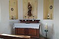 Kleiner Kirchenaltar einer Kapelle mit Marienstatue, Kerzen und Blumenschmuck vor weiß-gelb gestrichener Wand mit Ausfräsungen in der Wand mit gemalten Verzierungen darin. Im Vordergrund eine Gebetsbank aus Holz.