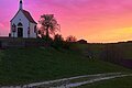 Links im Bild eine kleine Kapelle auf einem Hügel mit einem Weg unten am Hügel, umgeben von Bäumen und Weide. Sonnenuntergang mit pink-orangefarbenem Himmel im Hintergrund.