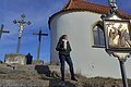 Frau steht auf einer Steintreppe neben einer Kapelle mit drei Kreuzen im Hintergrund und einem religiösem Bild auf einer Tafel, bei blauem Himmel.