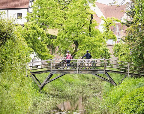 Zwei Personen mit Fahrrädern stehen auf einer Holzbrücke über der kleinen Wörnitz, umgeben von Bäumen und Altstadthäusern Donauwörths.