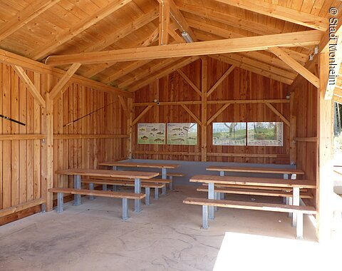 Überdachte Holzhütte mit Holzbänken und Tischen links und rechts an der Wand, an der Rückwand Infotafeln mit Fischabbildungen zu heimischen Fischarten angebracht.