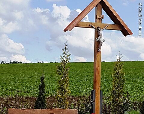 Flurkreuz am Wegesrand zwischen Monheim und Warching Holzkreuz mit überdachter Christusfigur mit bepflanztem Beet darunter, daneben eine Holzbank. Im Hintergrund ein Feld und blauer Himmel.