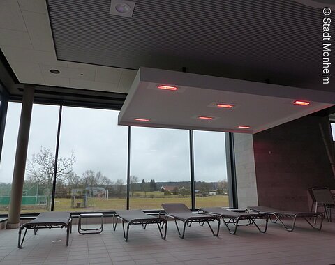 Innenraum eines Hallenbades mit Liegestühlen vor großen Fenstern und einer Deckenleuchte mit roten Lichtern.