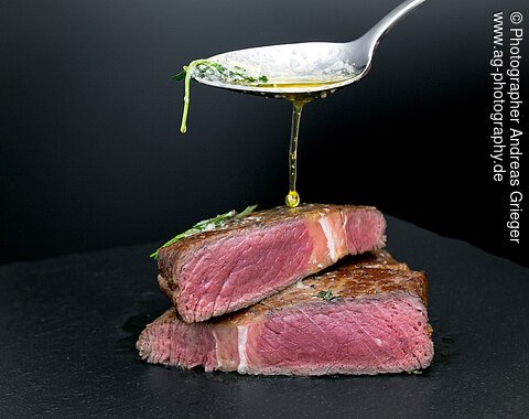 Ein Löffel gießt Öl über zwei aufeinander gestapelte, halbierte Rindersteaks auf einem schwarzen Hintergrund.