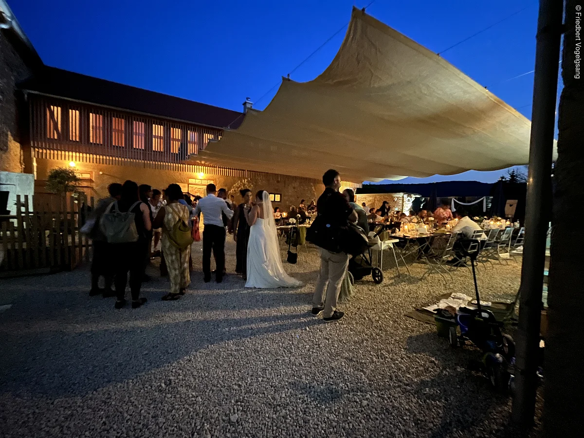 Menschen bei Abendveranstaltung unter großem Sonnensegel vor beleuchtetem Gebäude auf Kiesplatz.