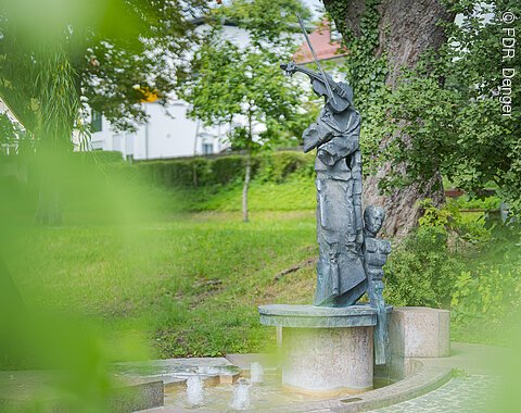 Statue einer Person mit Geige und Kind, umgeben von Bäumen und Grünfläche.