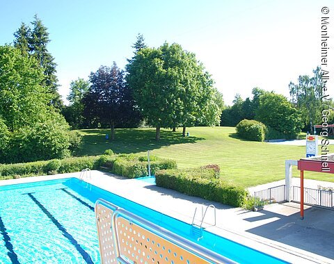 Außenbereich eines Schwimmbads mit Becken, Liegewiese, Bäumen und Uhr an einem sonnigen Tag.