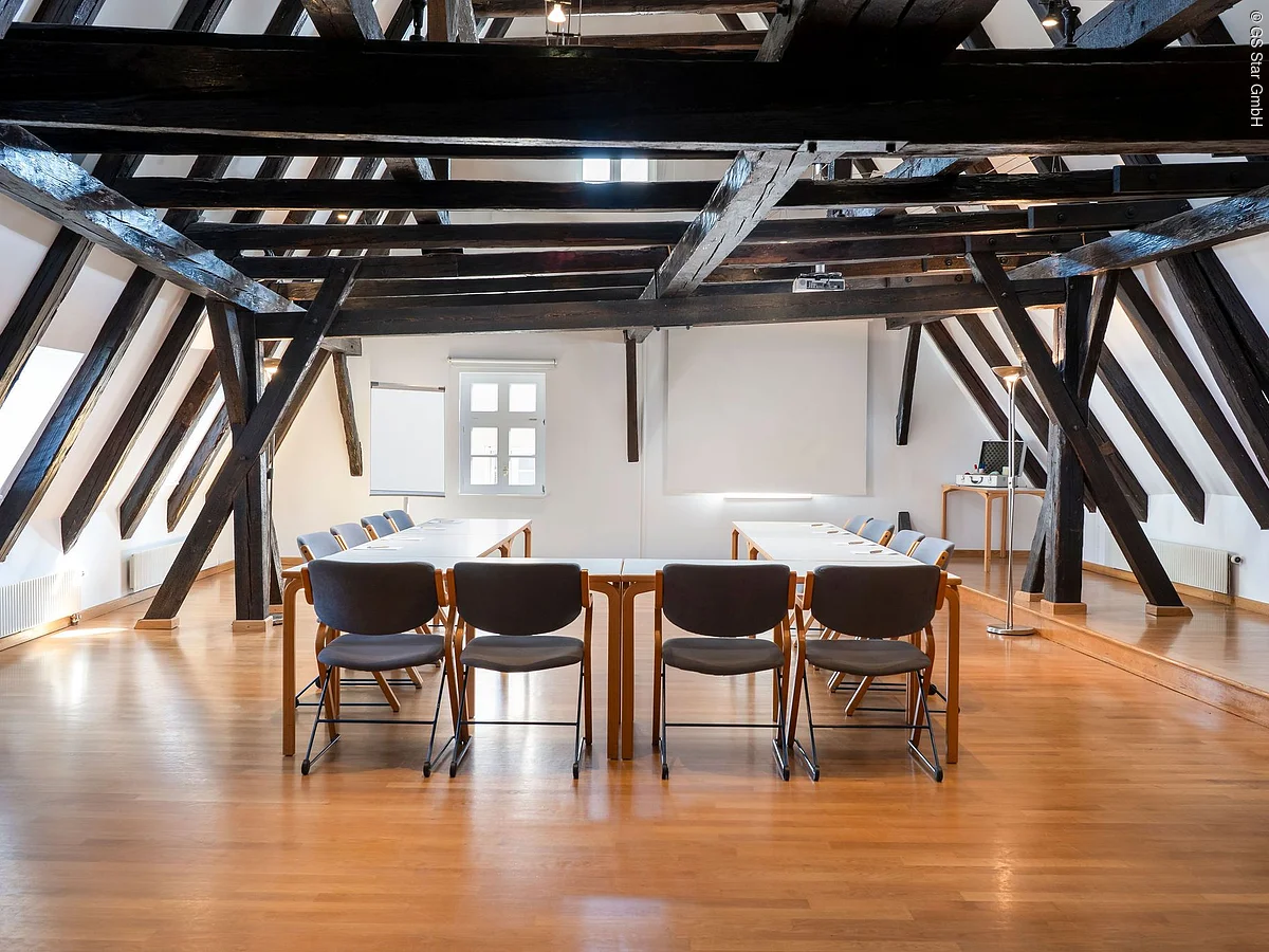 Konferenzraum mit Holzboden, dunklen Holzbalken und Tischen in U-Form mit Stühlen im Dachgeschoss.