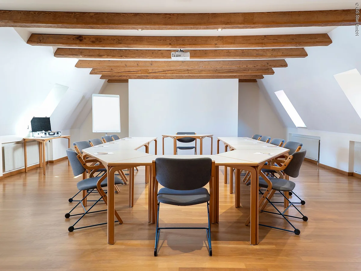 Konferenzraum mit U-förmigen Tischen, grauen Stühlen, Holzdecke mit Balken und weißer Leinwand.