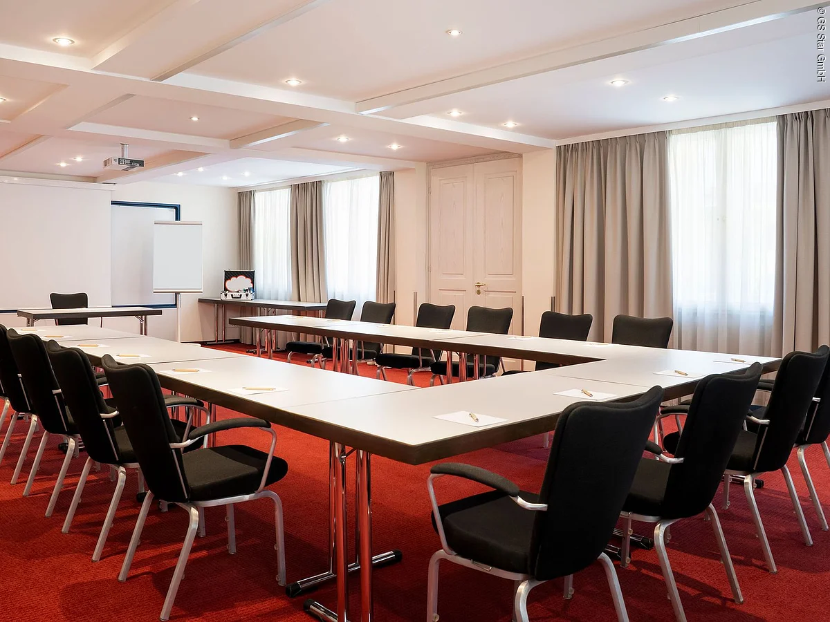 Konferenzraum mit U-förmigen Tischen, schwarzen Stühlen, rotem Teppich und weißem Flipchart vor Projektionsleinwand.