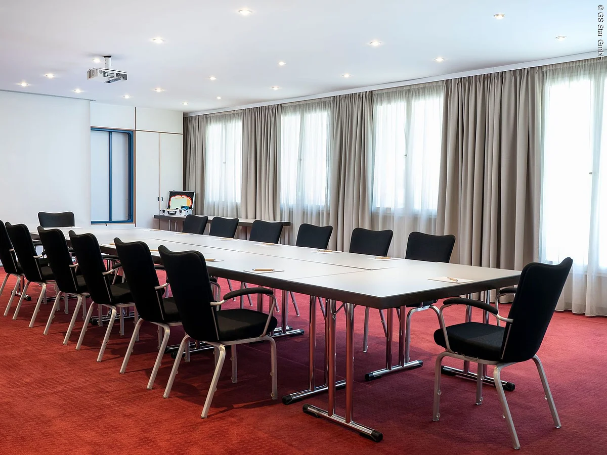 Konferenzraum mit langem Tisch, schwarzen Stühlen, roten Teppichboden und großen Fenstern mit Vorhängen.