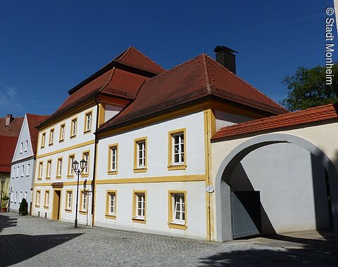 Pfarr- und Stadtbücherei Monheim Historisches weißes Gebäude, die Pfarr- und Stadtbücherei in Monheim, mit gelben Fensterrahmen und rotem Dach, daneben ein Torbogen auf gepflasterter Straße. Links vom Gebäude weitere Häuser in blau und grün.