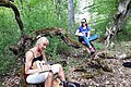 B+ Zentrum Blossenau - Uke-Wanderung Zwei Frauen mit Ukulelen sitzen im Wald auf einem moosbedeckten, gebogenen Baumstamm, eine schreibt.