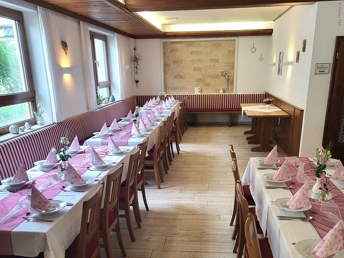 Gastraum mit zwei langen Tischen, gedeckt mit weißen Tüchern, rosa Servietten und Holzmöbeln.