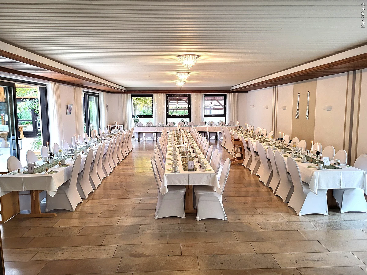 Großer Raum mit drei langen, festlich gedeckten Tischen und weißen Stuhlbezügen, Fenster und Türen links.