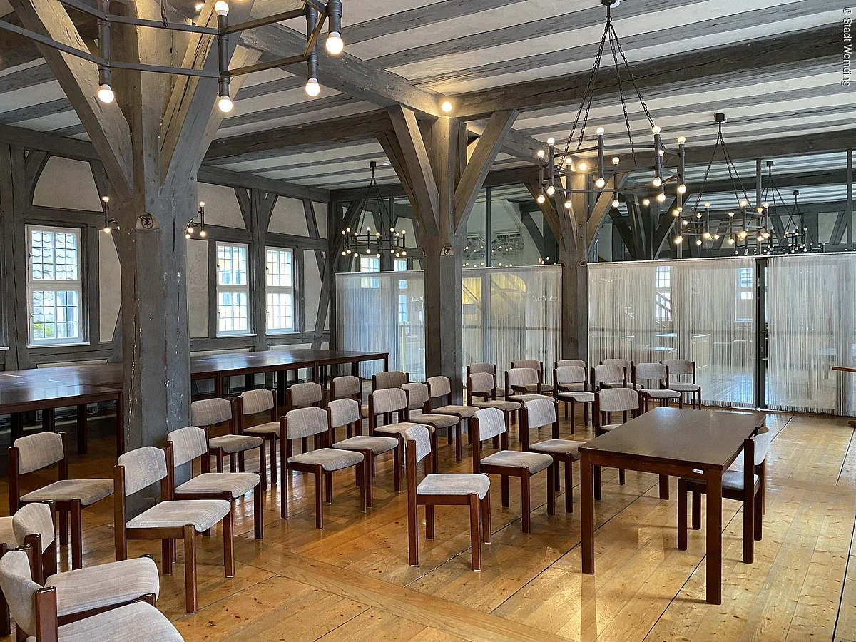 Großer Raum mit Holzboden, mehreren Reihen gepolsterter Stühle und Holztischen unter sichtbaren Holzbalken und Deckenlampen.