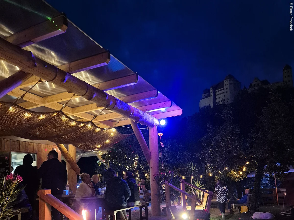 Abendliche Gartenbar mit Holzdach, Sitzbänken, Lichterketten und Menschen, im Hintergrund beleuchtetes Schloss Harburg auf Hügel.