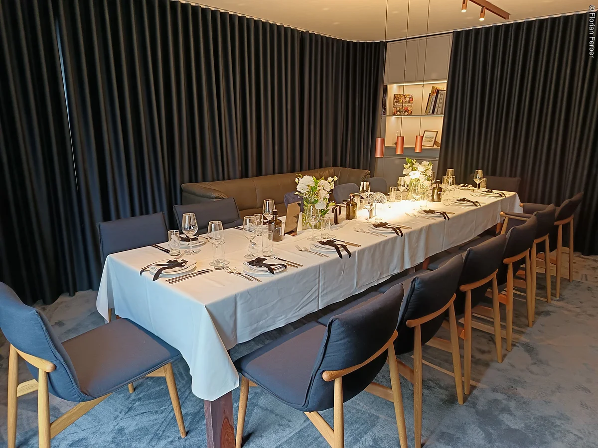 Langer gedeckter Esstisch mit weißen Tischdecken, Stühlen und dunklen Vorhängen im Hintergrund.